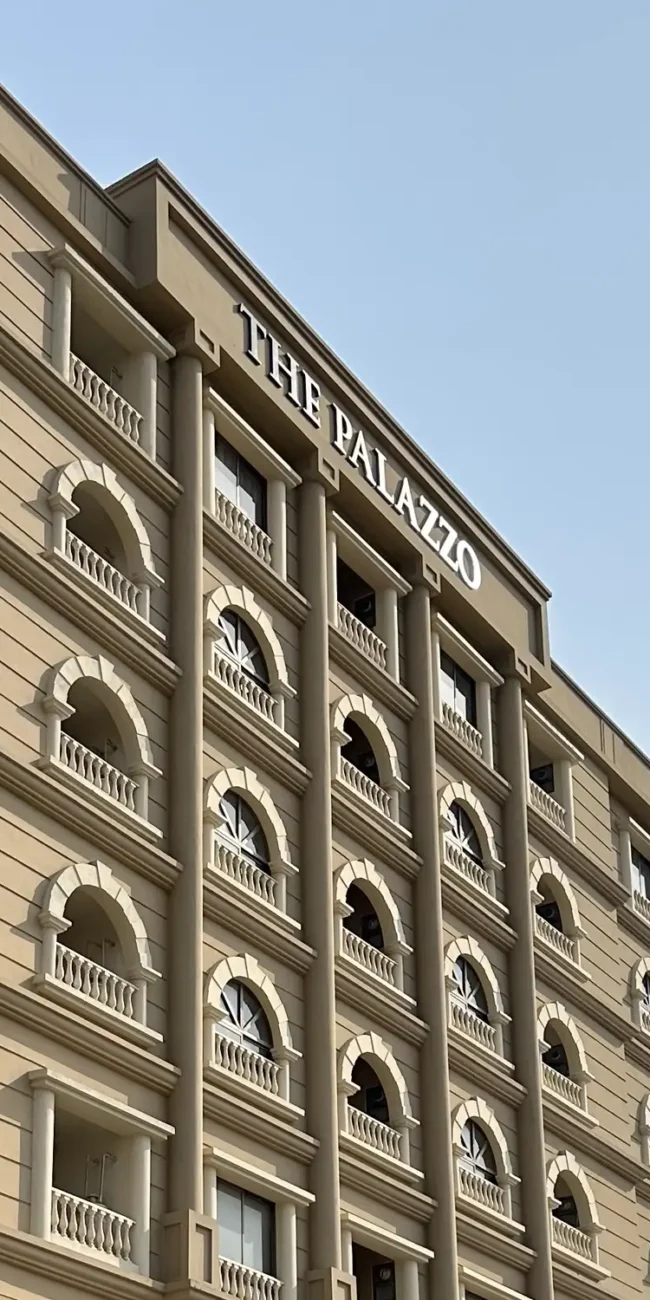 Angled view of The Palazzo commercial building design located in Gulberg Green, Rawalpindi/Islamabad, showcasing its light brown facade, arched balconies with decorative railings, and prominent "THE PALAZZO" signage at the top against a clear sky.