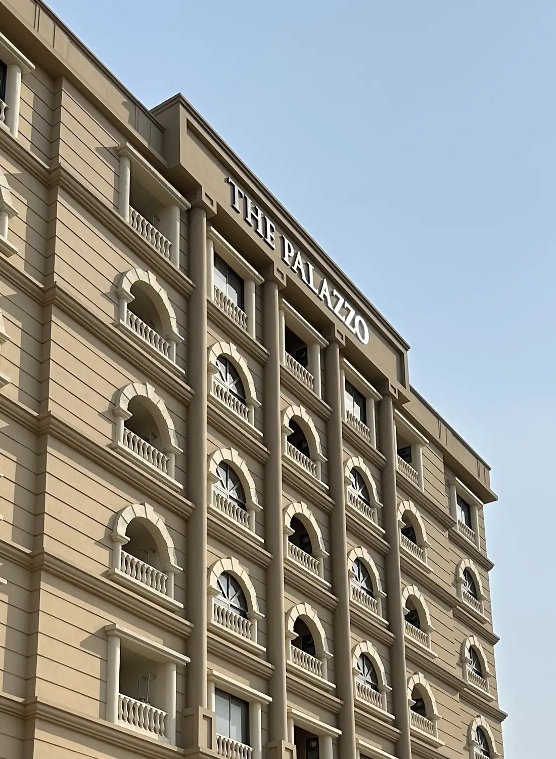 Angled view of The Palazzo commercial building design located in Gulberg Green, Rawalpindi/Islamabad, showcasing its light brown facade, arched balconies with decorative railings, and prominent "THE PALAZZO" signage at the top against a clear sky.