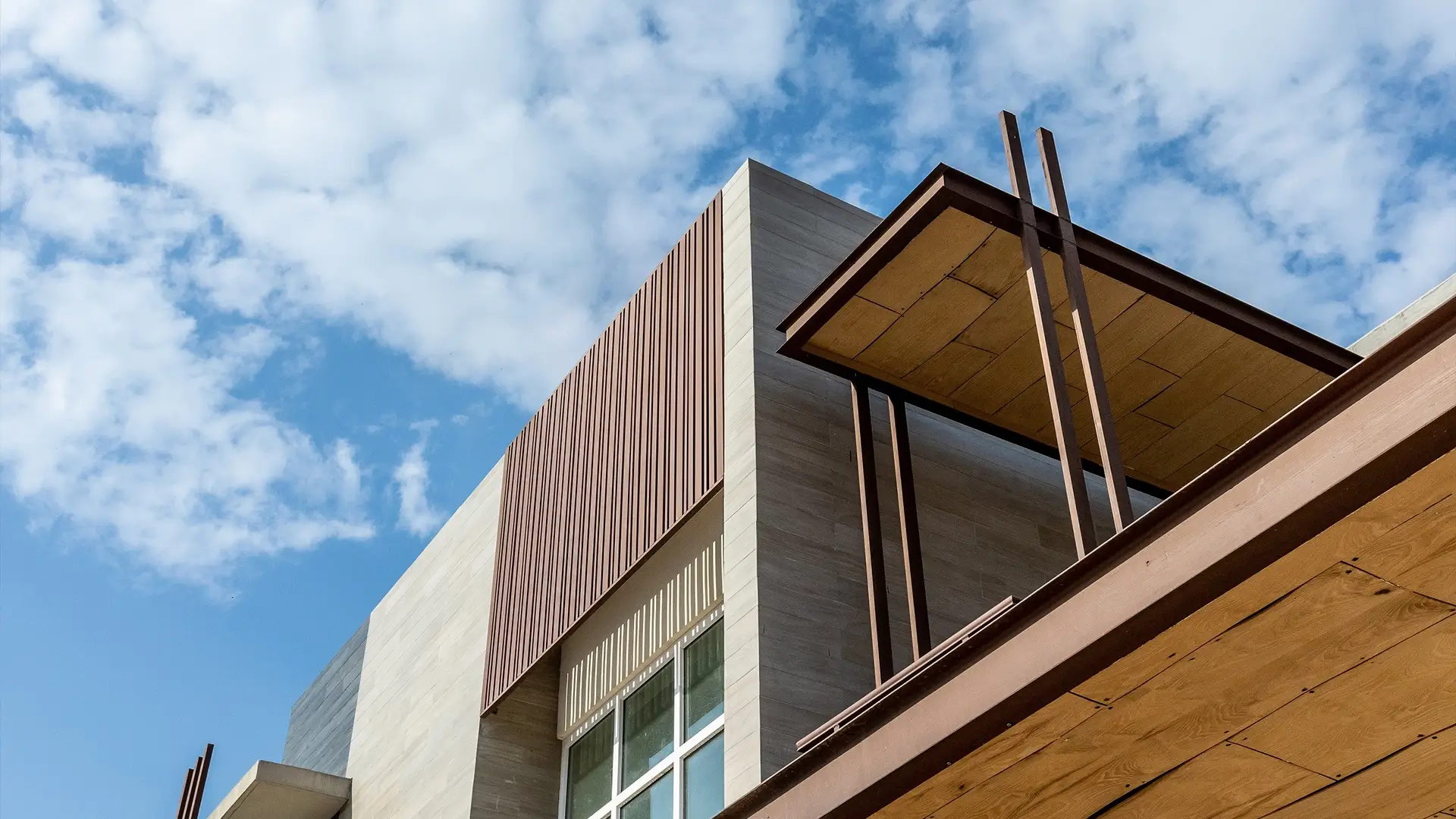 Modern house exterior showing a section of the upper facade with light-colored stone cladding, vertical brown slats, large windows, and a cantilevered roof detail against a cloudy sky.