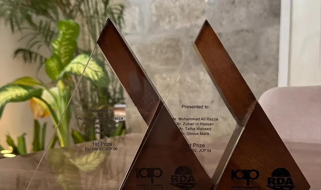 Two triangular glass and wood trophies are displayed on a reflective surface. Engravings on the trophies indicate "Presented to Ar. Muhammad Ali Razza & Ar. Talha Waheed" and "1st Prize" for the "Urban Monument Design Challenge for Slip Rd 01, JCP 06" and "Kashmir Rd 02, JCP 04." Logos for IAP and RDA are also visible on the trophies.