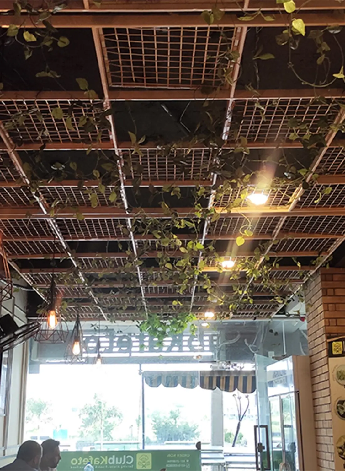 Interior view of a casual dining cafe in Islamabad, featuring a unique ceiling design with wooden lattice, integrated lighting fixtures, and hanging greenery, creating a warm and inviting ambiance. This small cafe in J7 Mall Islamabad offers a distinctive dining experience.