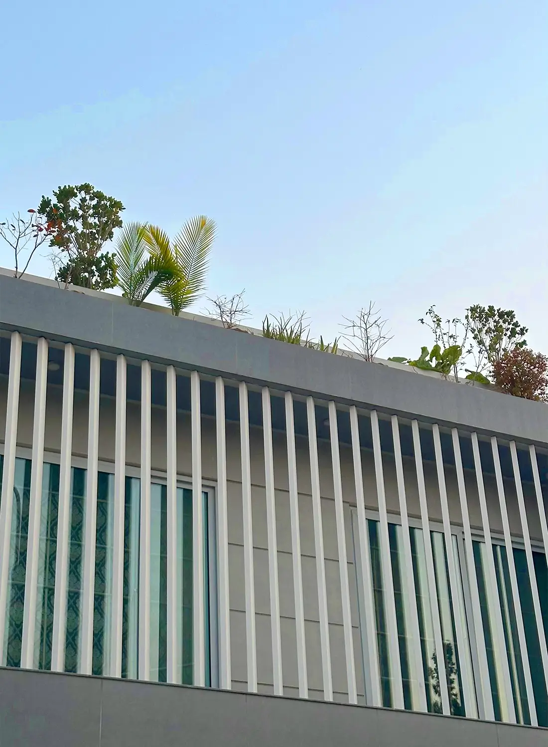 This image shows the upper section of a modern house in Islamabad, featuring a design with prominent white vertical louvers on the first floor. Above the louvers, a green roof with various small trees and plants is visible against a clear sky. This architectural detail, part of a residential project by Primarc Studio, highlights a contemporary approach to building design in Islamabad.