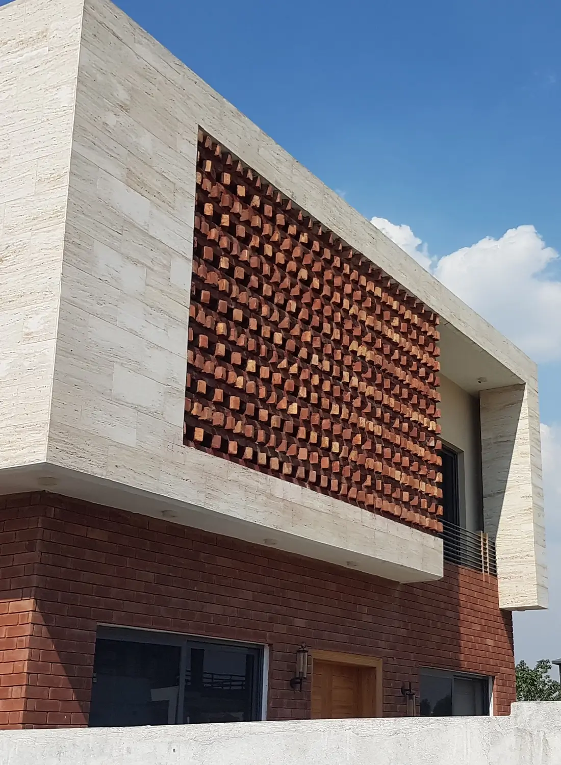 Modern house exterior in DHA Phase 2, Islamabad, featuring a prominent upper section with light-colored travertine cladding and a large decorative screen made of projecting red bricks arranged in a pattern. The ground floor is clad in darker red bricks, with dark-framed windows and a wooden door visible. A concrete boundary wall is in the foreground, and a clear blue sky with some clouds is in the background.