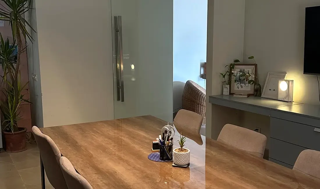 A modern and spacious conference room features a large wooden table surrounded by comfortable chairs. A long, rectangular pendant light illuminates the table. Plants and a glimpse into an adjacent office space with a desk and chair are visible in the background.