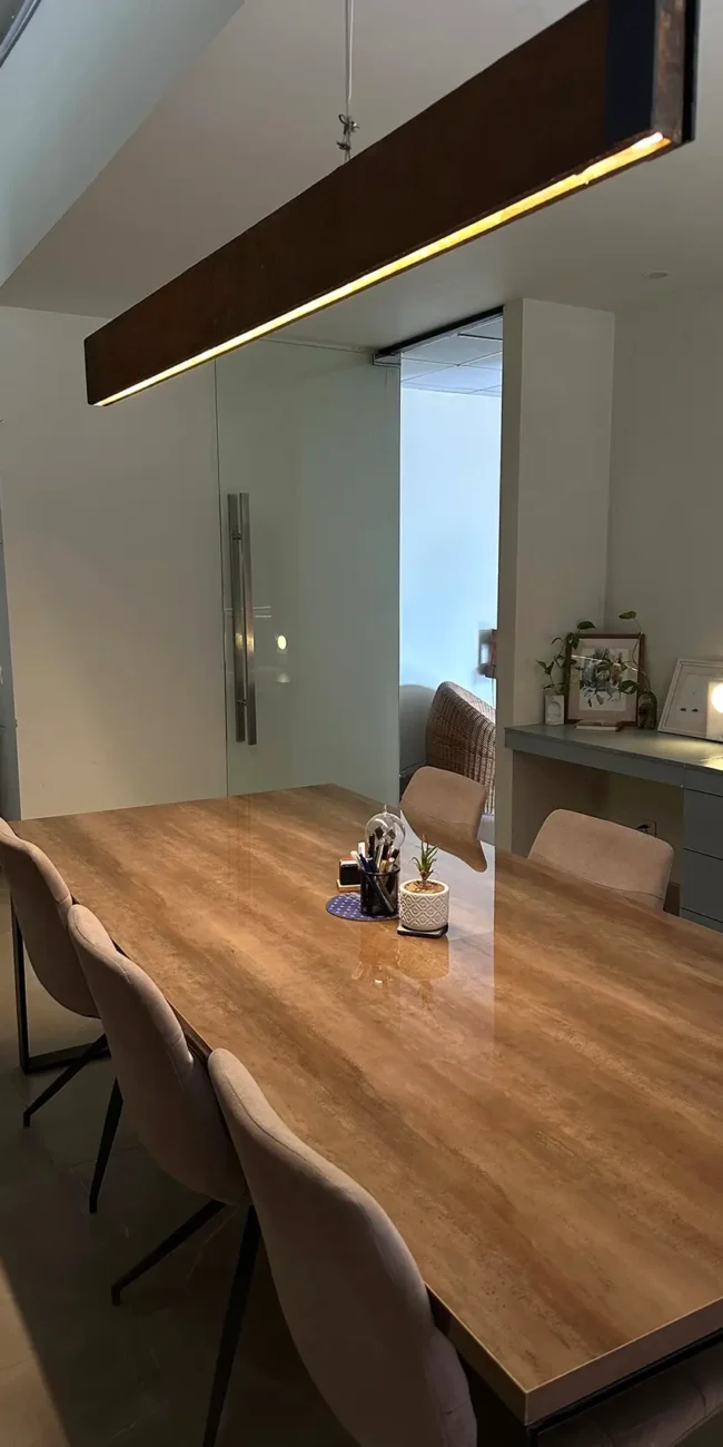 A modern and spacious conference room features a large wooden table surrounded by comfortable chairs. A long, rectangular pendant light illuminates the table. Plants and a glimpse into an adjacent office space with a desk and chair are visible in the background.
