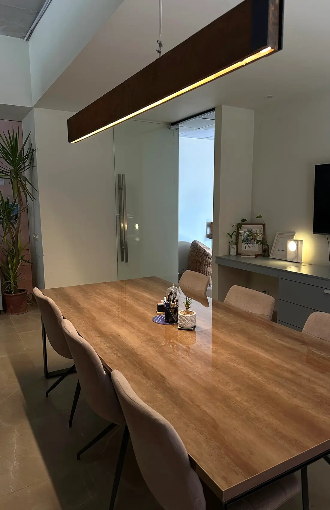 A modern and spacious conference room features a large wooden table surrounded by comfortable chairs. A long, rectangular pendant light illuminates the table. Plants and a glimpse into an adjacent office space with a desk and chair are visible in the background.