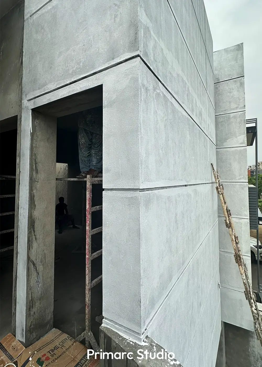 Exterior view of a minimalist house under construction, showing a concrete facade with horizontal bands and a large entrance opening.