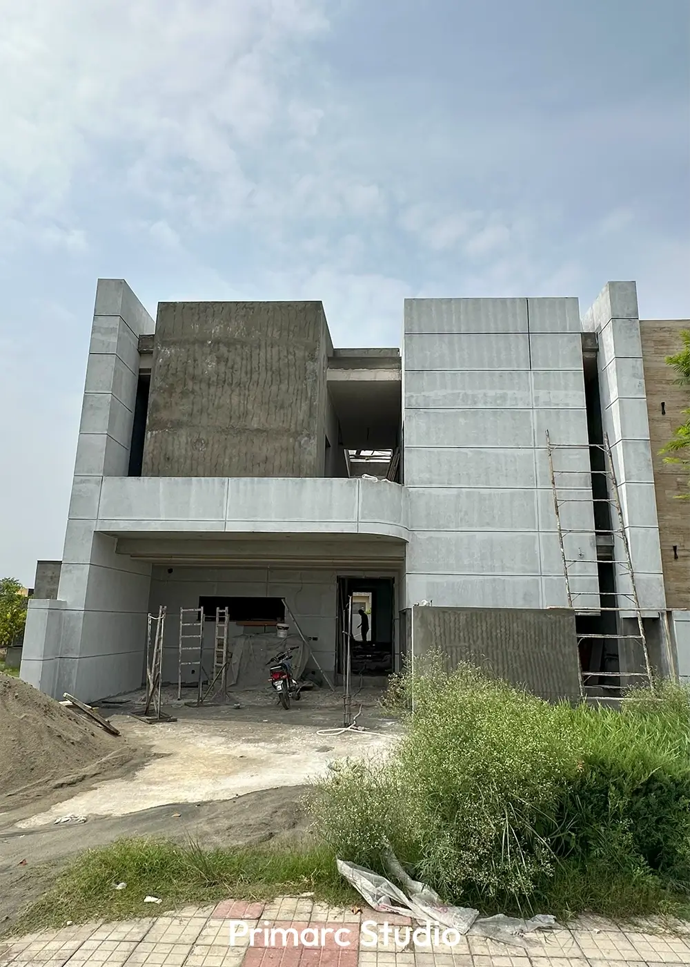 Front exterior view of a minimalist house under construction, with concrete facade, a car porch, and surrounding landscape.