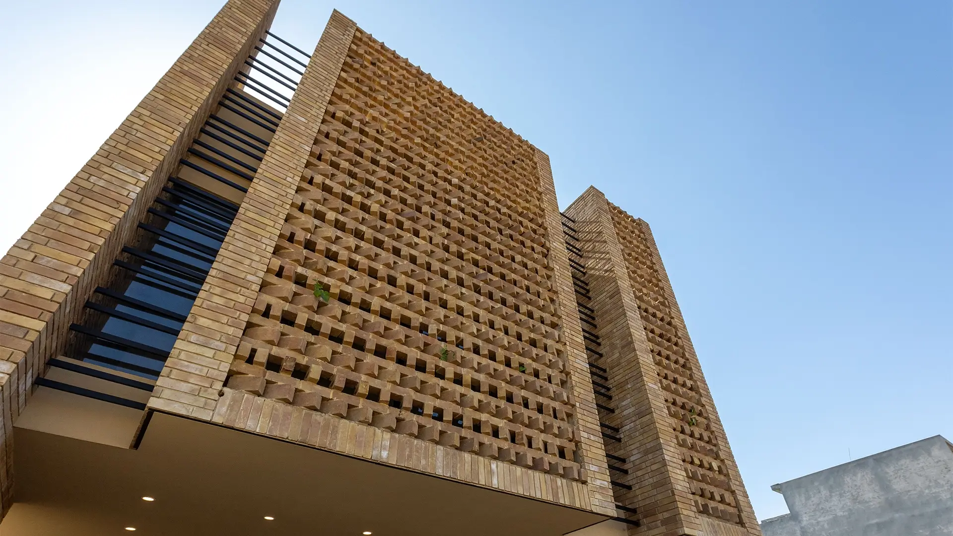 Low-angle view of a modern building's facade, featuring an intricate, three-dimensional screen of honey-colored brickwork against a clear blue sky.
